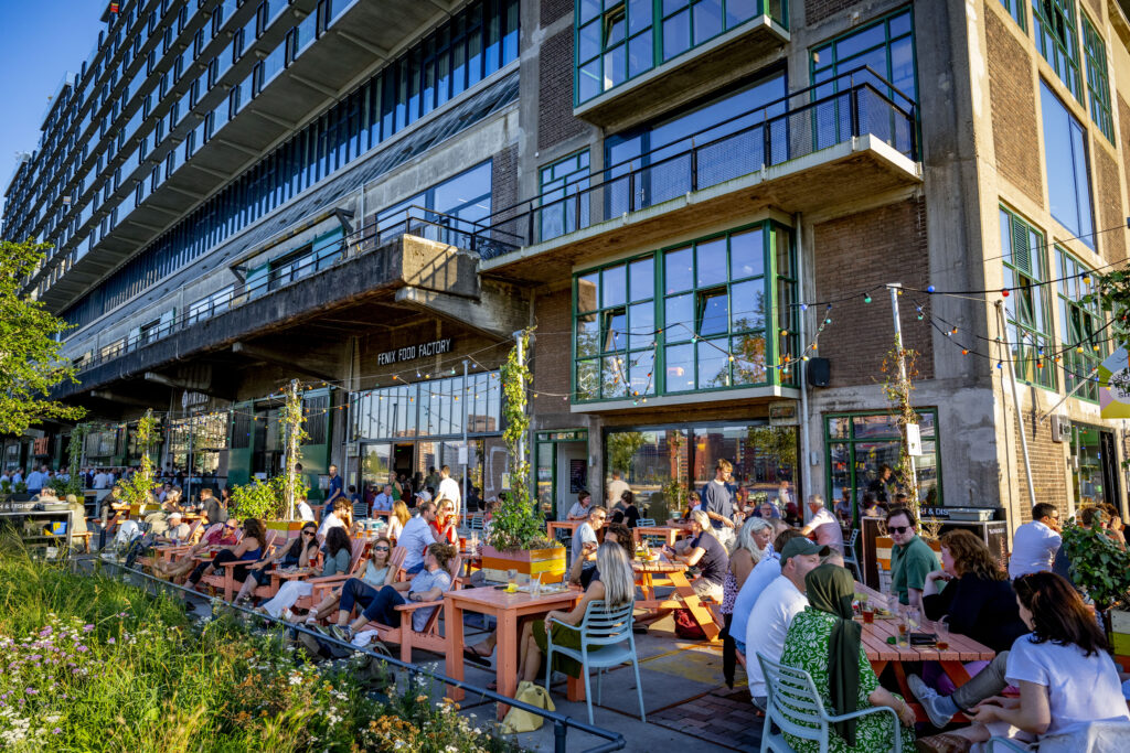 Een foto van mensen die genieten van een zomeravond op de Kop van Zuid, bij de Fenix Food Factory - een culinair centrum met een foodhall, bijzondere brouwerij en kraampjes voor vlees, groenten en zuivel.